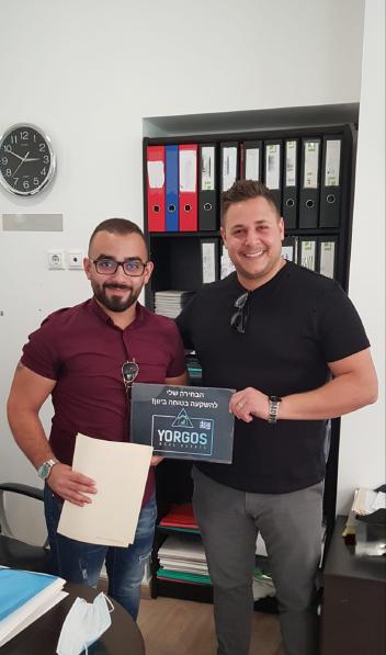 Happy client of Yorgos Real Estate company with big smile holding the documents of their newly purchased property, standing next to the company owner Mr. Yorgos.